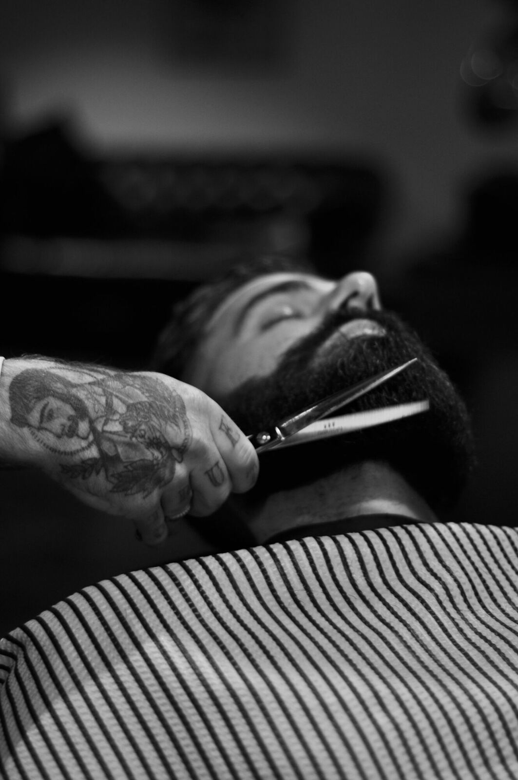 A tattooed barber's hand holds scissors near the beard of a reclining man wearing a striped cape.