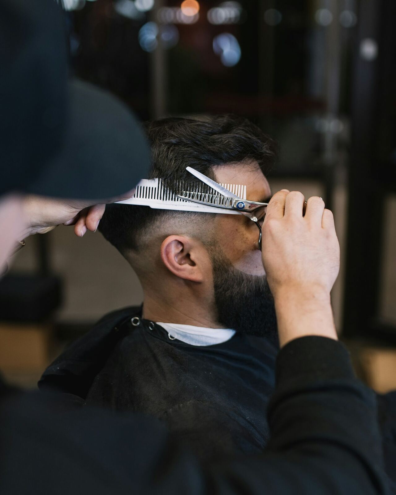 A barber uses scissors and a comb to cut the hair of a seated man wearing a cape.