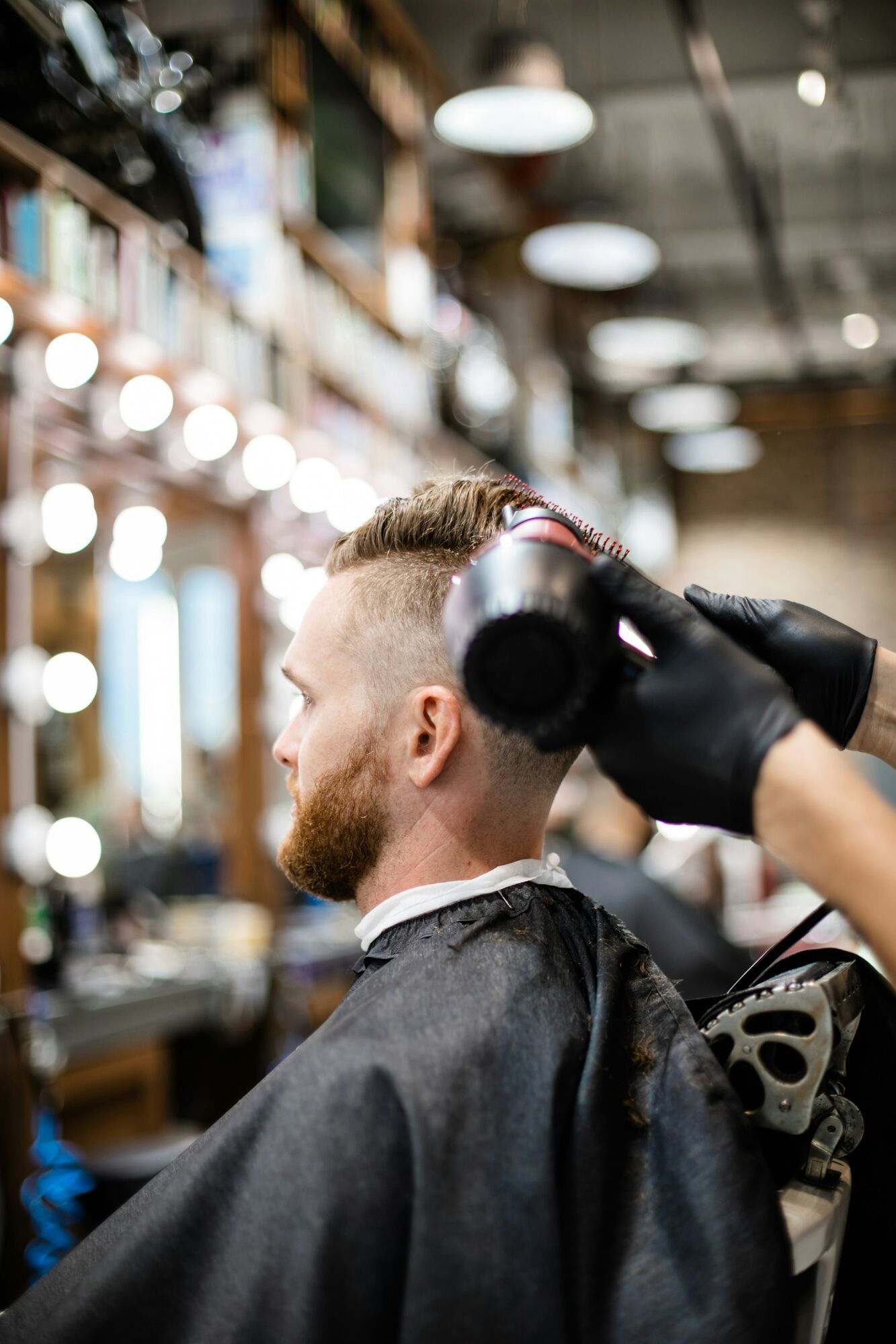 A barber wearing black gloves uses a hair dryer on a bearded man's hair in a barbershop.